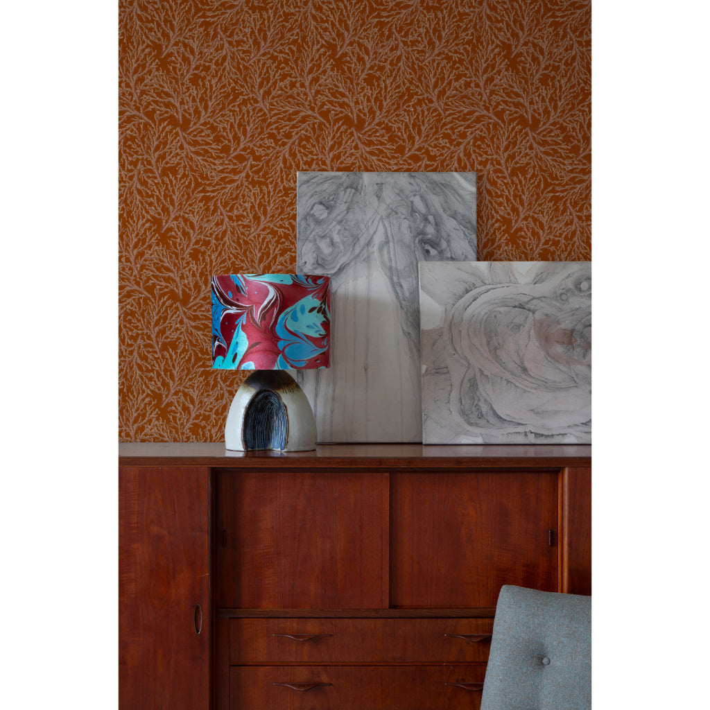 A styled interior scene showing a wooden sideboard with a colourful marbled lamp, two abstract canvases leaning against a patterned orange wall, and part of a grey upholstered chair in the foreground.