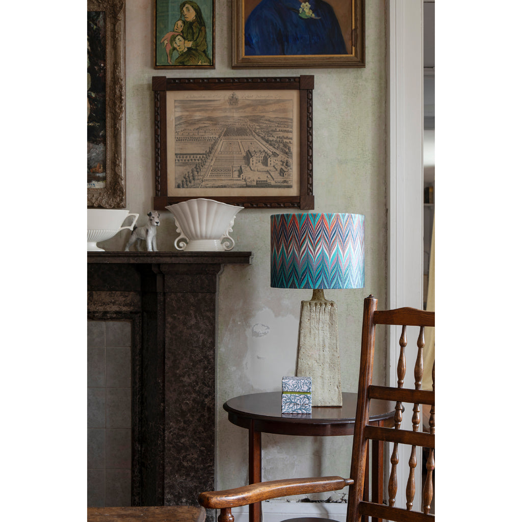 A styled interior scene featuring a table lamp with a colorful chevron-patterned blue and orange shade, set next to a wooden chair in front of a dark fireplace. Framed art hangs on the wall above.