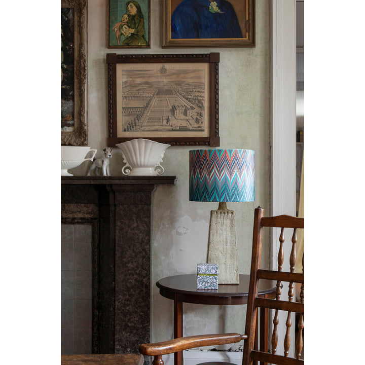 A styled interior scene featuring a table lamp with a colorful chevron-patterned blue and orange shade, set next to a wooden chair in front of a dark fireplace. Framed art hangs on the wall above.