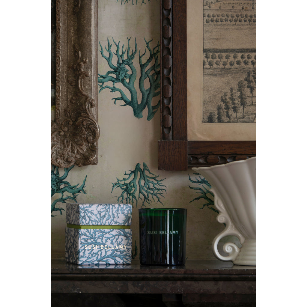 A styled interior scene showing a dark green glass candle jar and a patterned gift box on a wooden console table, set against wallpaper with teal coral-like botanical illustrations and framed artwork.