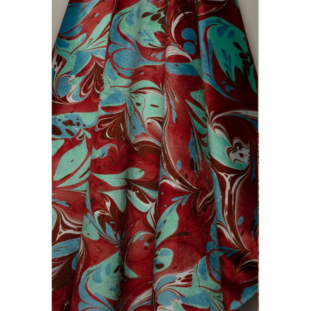 A close-up of a draped fabric with a marbled pattern in deep red, teal, brown, and white, shown against a plain off-white background.