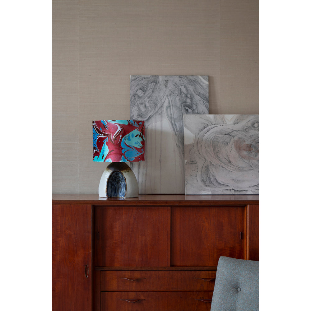 A drum lampshade featuring a marbled pattern of red, teal, brown, and white on a midcentury sideboard with art in the background