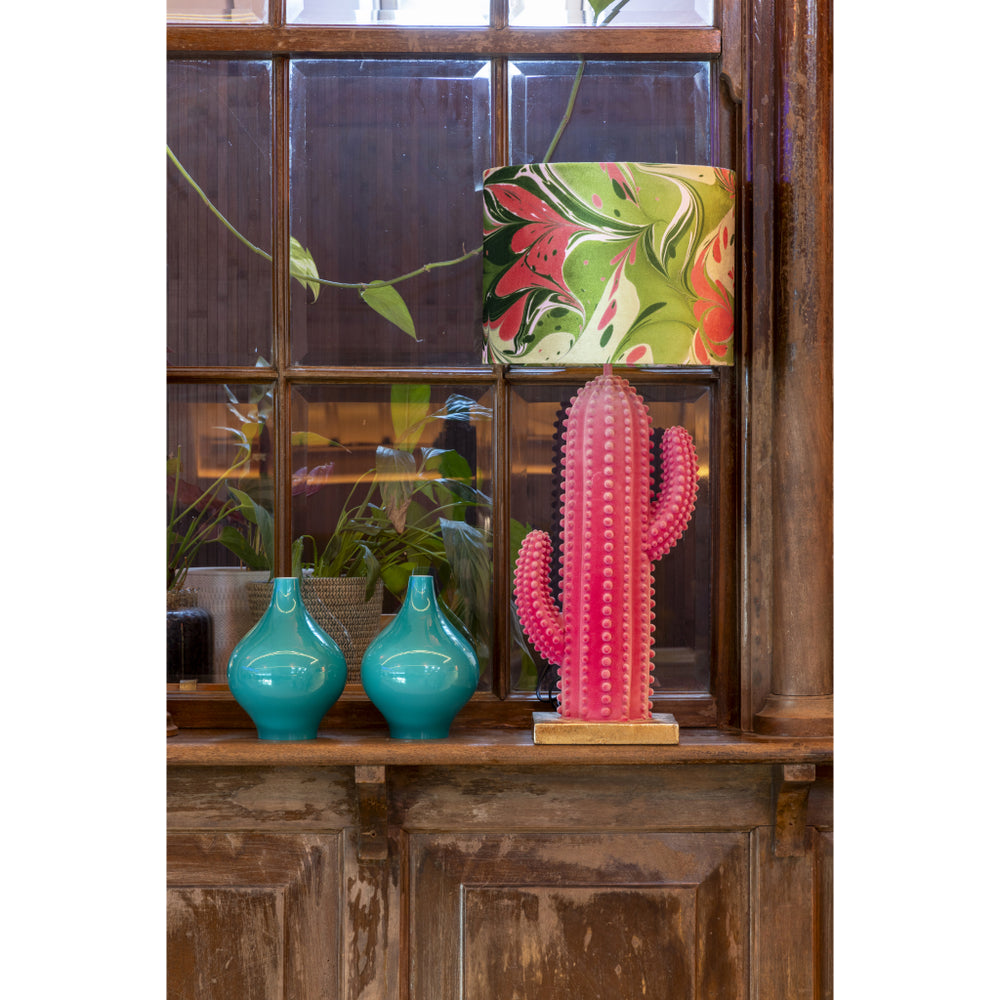 A decorative pink cactus lampbase with a colourful marbled pattern velvet lampshade sits on a rustic wooden shelf next to two turquoise vases in front of a dark wood window.