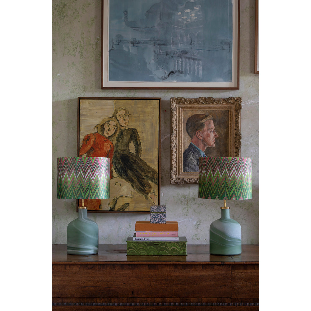 A styled interior scene showing two matching table lamps on a wooden console table, with framed artwork and books on the wall behind them. The lamps have swirled sage green ceramic bases and patterned drum shades with a chevron design in green, pink, and white.