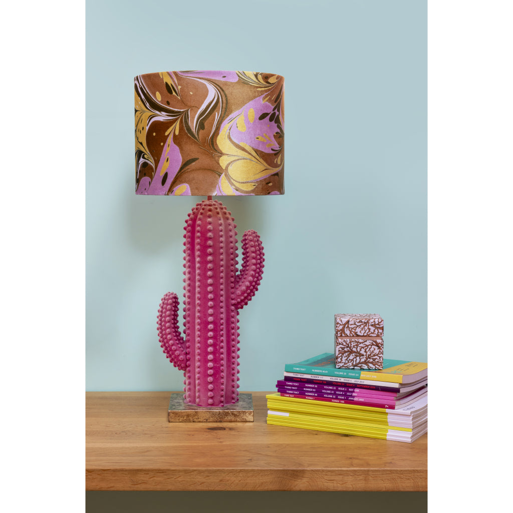 A table lamp with a pink cactus-shaped base and a marbled shade in brown, gold, and purple, styled on a wooden table next to a stack of colorful books against a light blue wall.