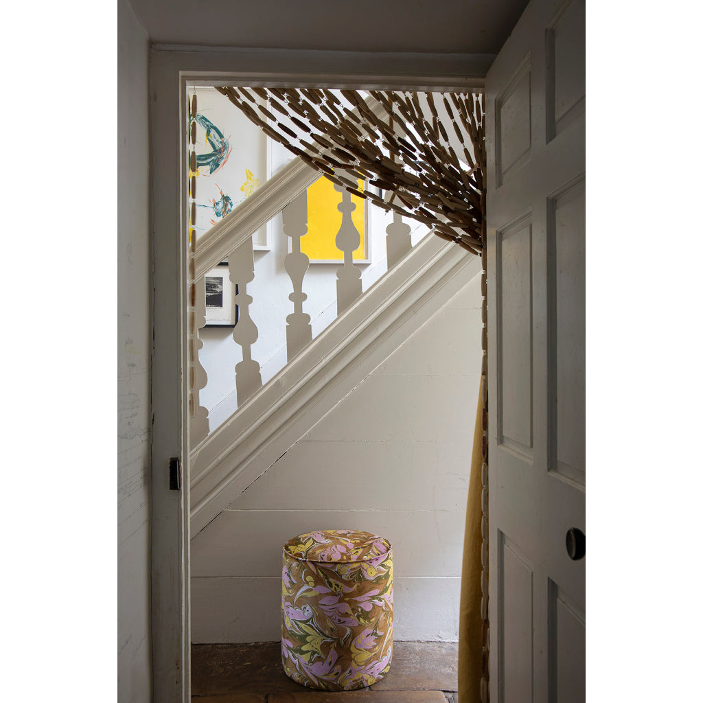 A cylindrical upholstered stool with a brown, lavender and yellow marbled pattern sits on a wooden floor in a doorway, looking onto a white staircase with a yellow wall art piece in the background