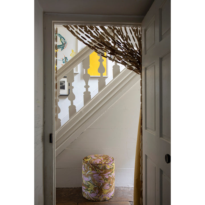 A cylindrical upholstered stool with a brown, lavender and yellow marbled pattern sits on a wooden floor in a doorway, looking onto a white staircase with a yellow wall art piece in the background