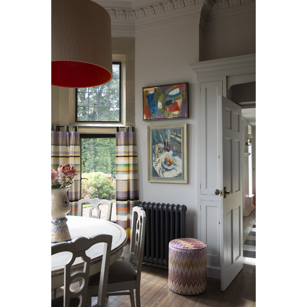 A styled interior dining room corner featuring two framed paintings on a light gray wall, a striped window curtain, a large drum pendant lamp with a red interior, wooden chairs, a round patterned ottoman, and a dark radiator.