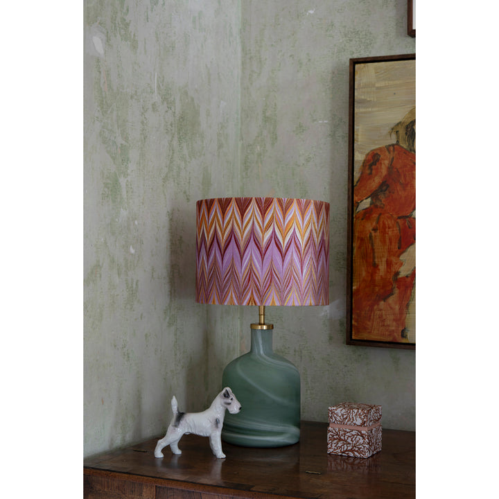 A table lamp with a green glass base and a pink, purple, and orange chevron-patterned shade, styled on a dark wooden table next to a small white dog figurine and a patterned box against a textured green wall. 