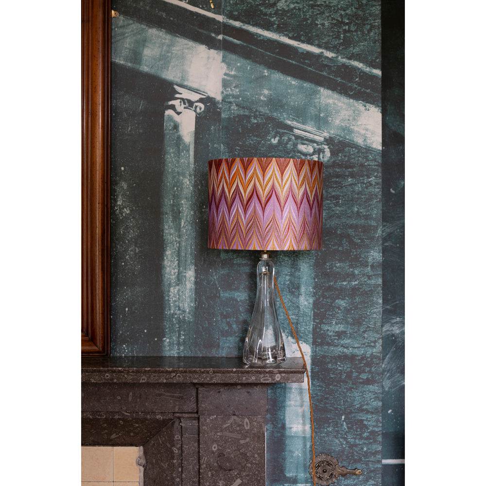A table lamp with a colourful zigzag patterned shade in pink, orange, and purple, set on a dark stone mantelpiece against a textured teal wall.