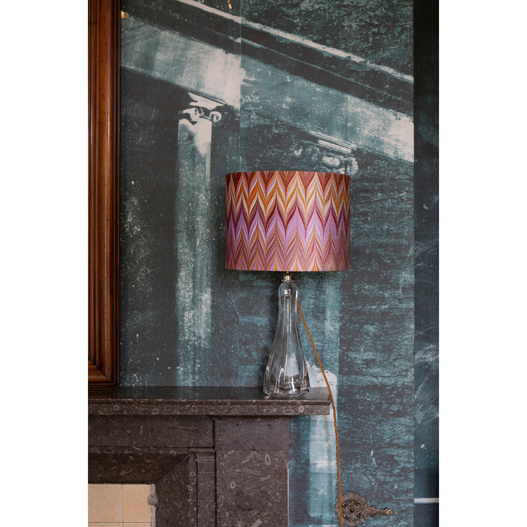 A table lamp with a colourful zigzag patterned shade in pink, orange, and purple, set on a dark stone mantelpiece against a textured teal wall.