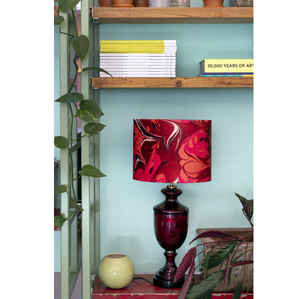 A table lamp with a glossy dark red glass base and a red, pink, and orange marbled shade, styled on a shelf next to a small vase and plants.