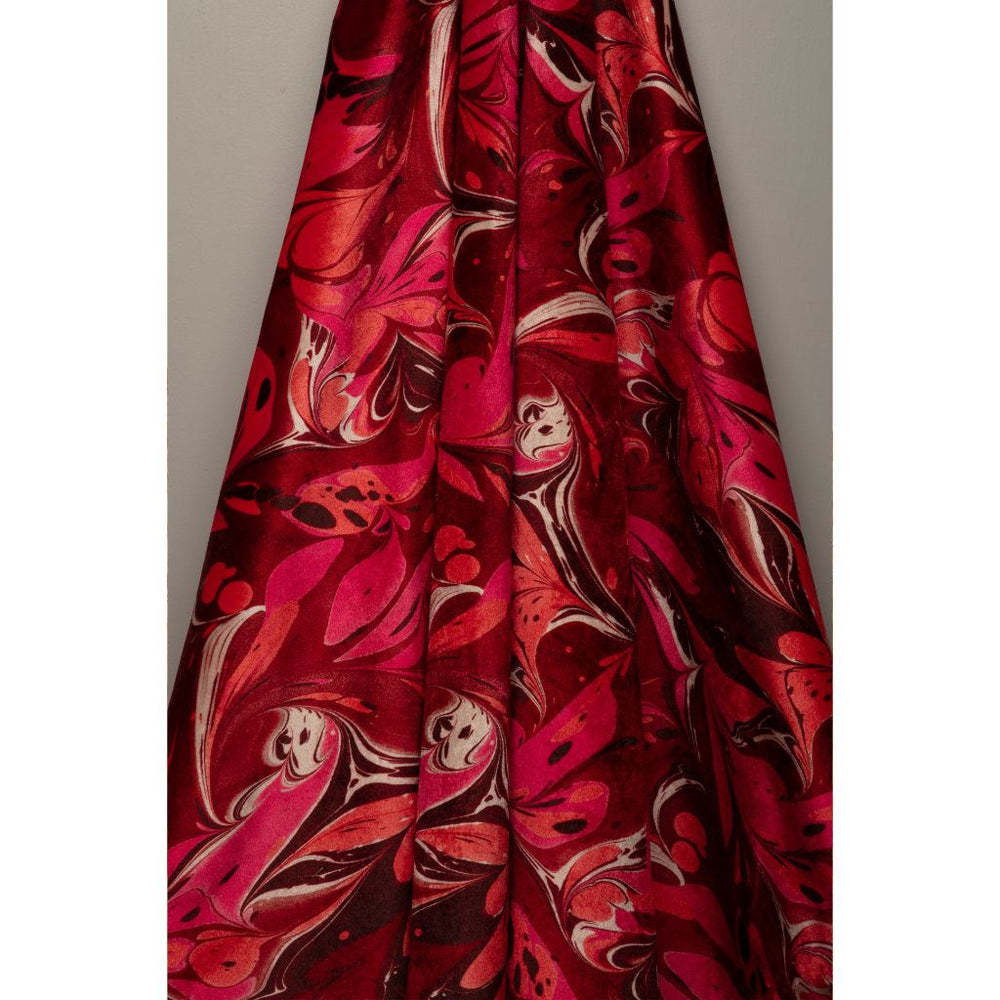 A close-up of a draped fabric with a bold abstract marbled pattern in vibrant red, magenta pink, and dark brown, set against a plain gray background.