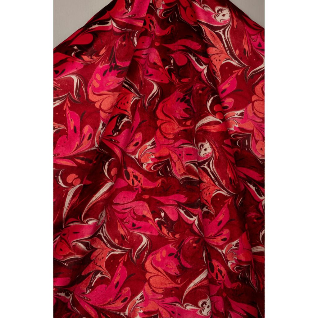 A close-up of draped fabric with an abstract marbled pattern in deep red, bright pink, coral, and cream swirls.
