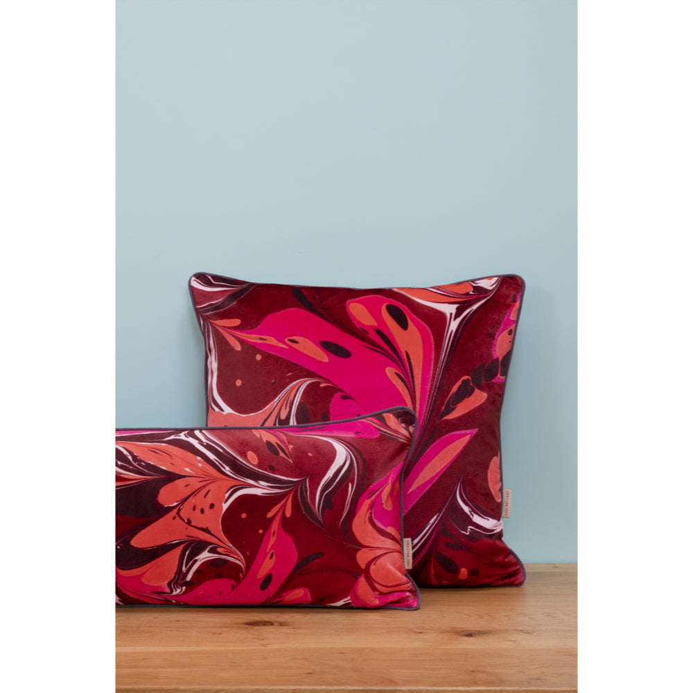 Two decorative throw pillows with a vibrant red, pink, and orange abstract marbled pattern, displayed on a light wood surface against a pale blue wall.
