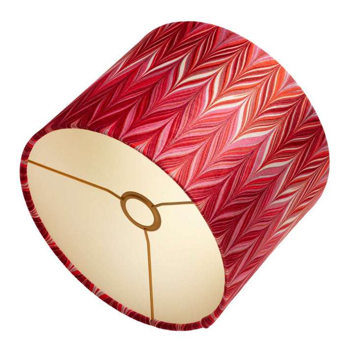 A cylindrical lampshade featuring a red and pink chevron pattern on the exterior and a cream-colored interior with a gold-colored metal ring.