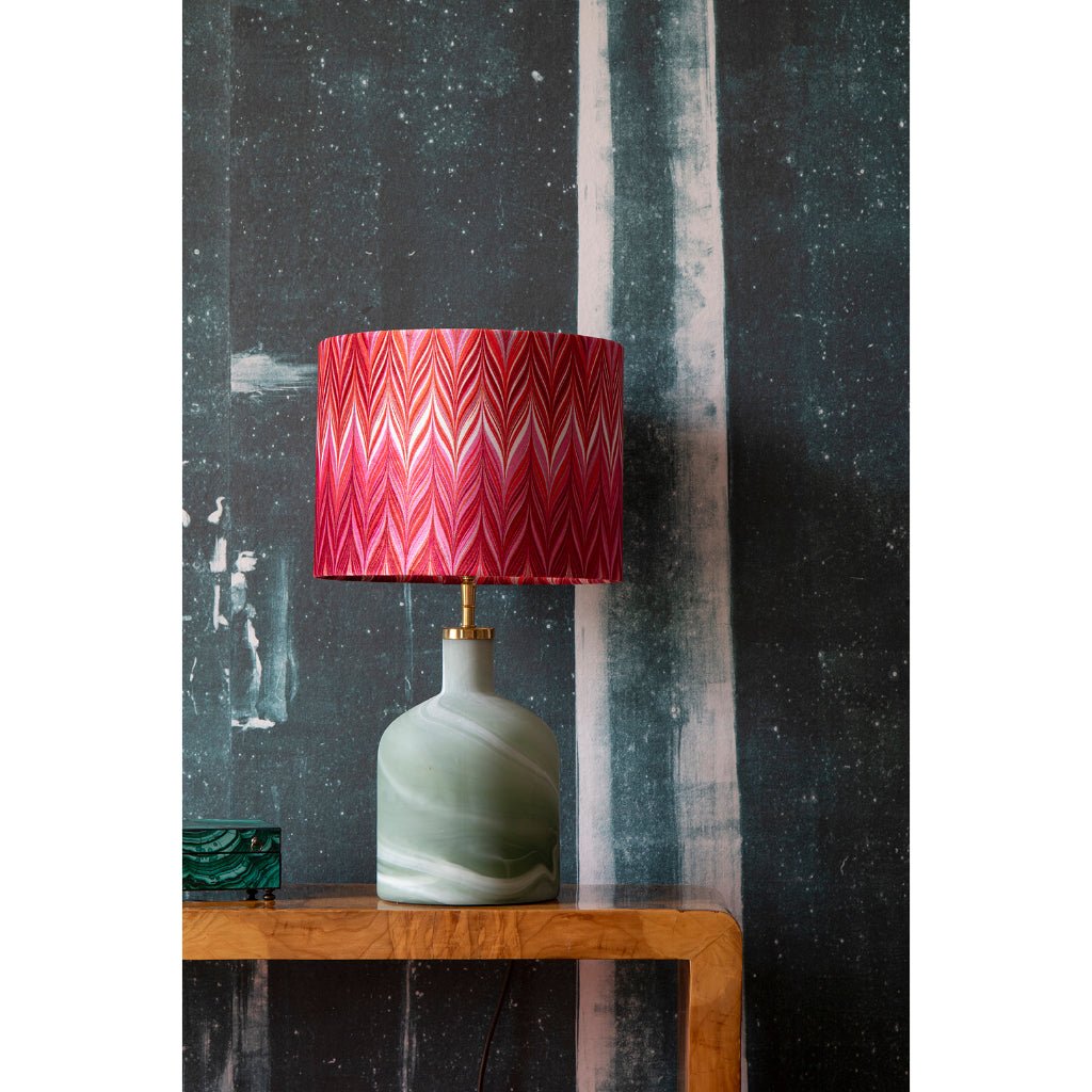 A table lamp with a marbled sage green ceramic base and a vibrant red and pink chevron-patterned velvet lampshade, displayed on a wooden side table against a dark textured wall.