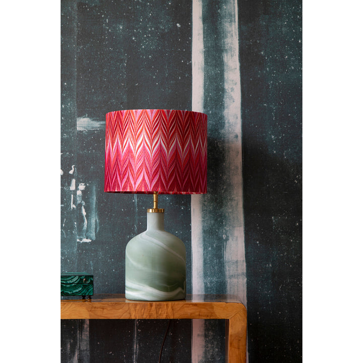 A table lamp with a marbled sage green ceramic base and a vibrant red and pink chevron-patterned velvet lampshade, displayed on a wooden side table against a dark textured wall.