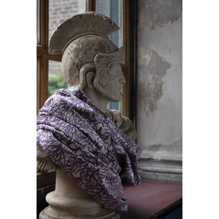 Statue of a warrior with a helmet and draped in a purple coral velvet, set against a textured wall