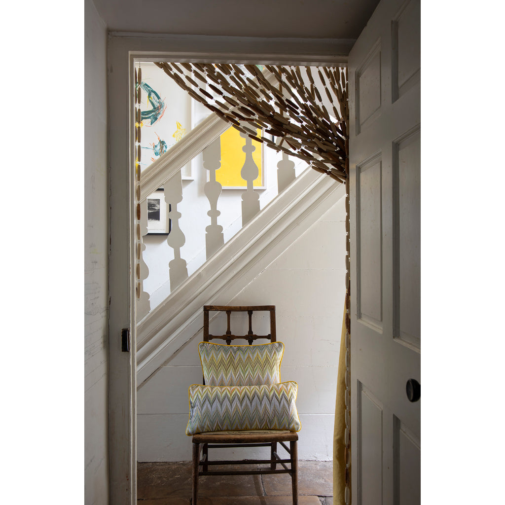 A rustic wooden chair with two patterned yellow, blue, and white chevron pillows sits in a doorway, looking up a white staircase. A decorative branch-like curtain hangs from the top of the stairs, and a white paneled door is visible on the right.