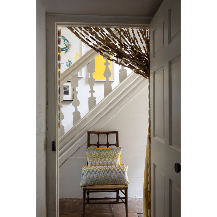 A rustic wooden chair with two patterned yellow, blue, and white chevron pillows sits in a doorway, looking up a white staircase. A decorative branch-like curtain hangs from the top of the stairs, and a white paneled door is visible on the right.