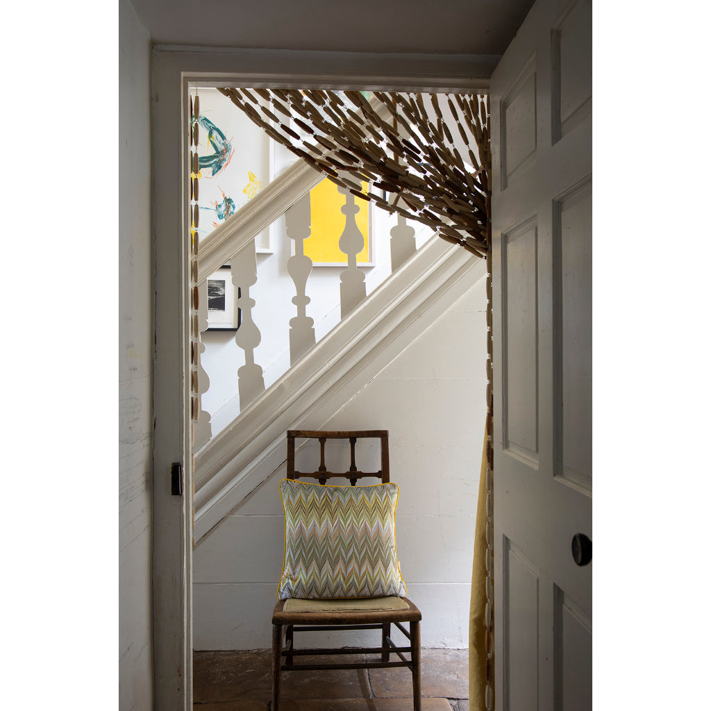 A rustic wooden chair with a yellow, green, and gray chevron-patterned throw pillow sits on a wooden floor in a doorway. Beyond the doorway is a white staircase with white balusters and a decorative branch-like curtain hanging near the top.