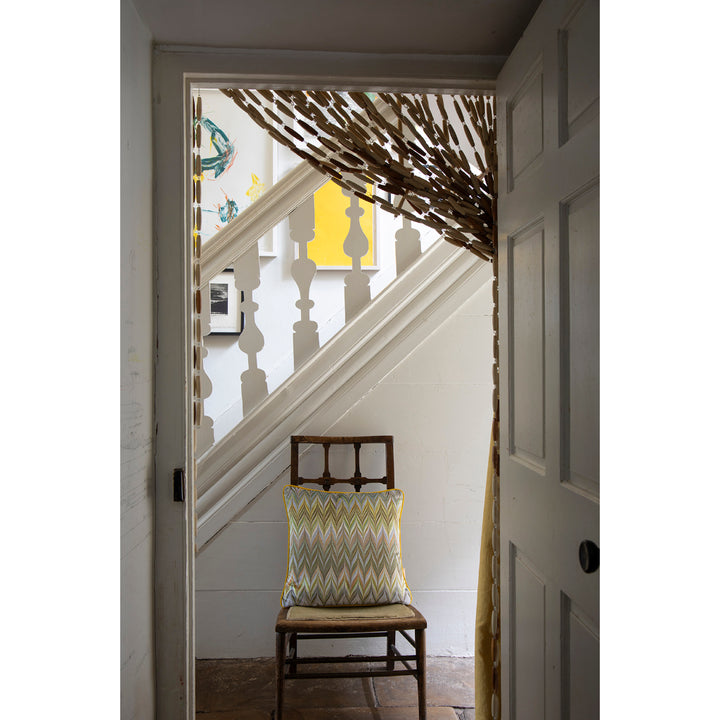 A rustic wooden chair with a yellow, green, and gray chevron-patterned throw pillow sits on a wooden floor in a doorway. Beyond the doorway is a white staircase with white balusters and a decorative branch-like curtain hanging near the top.