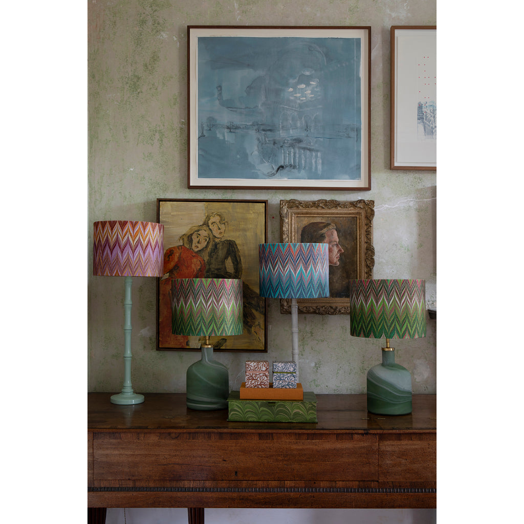 A styled interior scene showing a wooden console table with three table lamps featuring colorful zigzag-patterned drum shades and green ceramic bases, set against a textured wall with framed artwork.