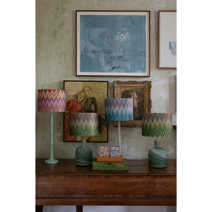A styled interior scene showing a wooden console table with three table lamps featuring colourful zigzag-patterned drum shades and green ceramic bases, set against a textured wall with framed artwork. 