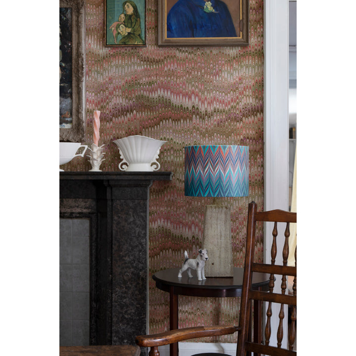 A styled interior scene featuring a table lamp with a blue, pink, and white chevron-patterned shade, set against a wall covered in matching patterned wallpaper. A dark fireplace mantel, framed paintings, a wooden chair, and a small side table are also visible.