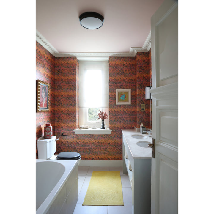 A narrow bathroom interior with orange, pink and red marbled pattern wallpaper, a white bathtub on the left, a double-sink vanity on the right, a yellow rug on the floor, and a window at the back.