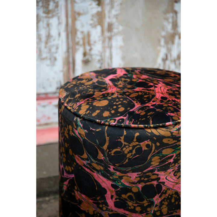 A close-up of a cylindrical drum stool upholstered in a marbled fabric with a black base, pink swirls, and brown/gold splatter patterns, set against a rustic weathered wall.
