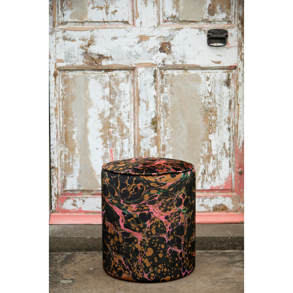 A close-up of a cylindrical drum stool upholstered in a marbled fabric with a black base, pink swirls, and brown/gold splatter patterns, set against a rustic weathered wall.