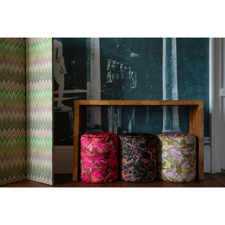 Three round velvet stools with marbled velvet fabric upholstery, patterns in bright pink, fuchsia pink, dark brown, black, brown, lilac and yellow, arranged in front of a wooden bench against a dark patterned wall
