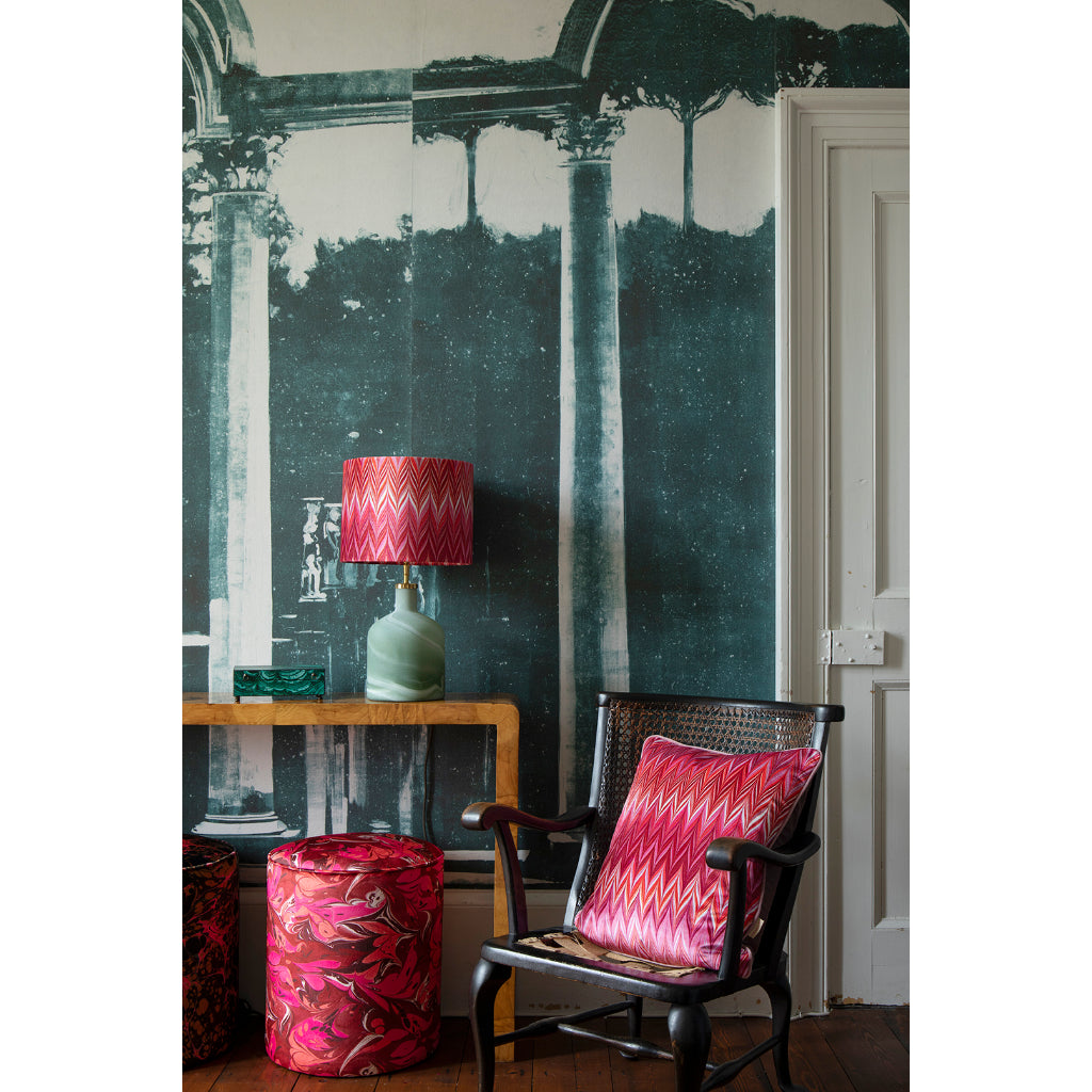 A styled interior corner featuring a dark wood armchair with a pink and red chevron-patterned throw pillow, a matching pink patterned velvet stool, and a table lamp with a pink chevron shade and pale green base, set against a large wall mural of classical columns.