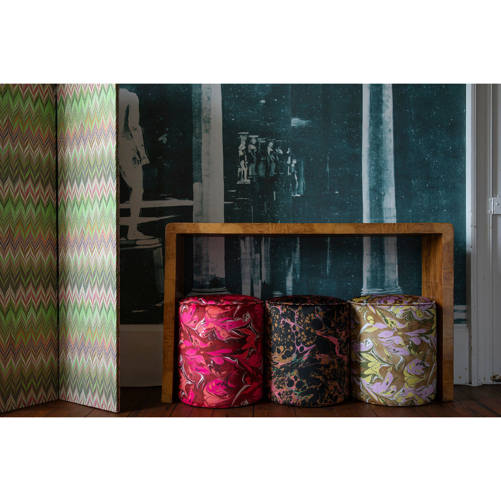 Three round velvet stools in various patterns in pink, gold, and green, arranged in front of a wooden bench against a dark patterned wall.