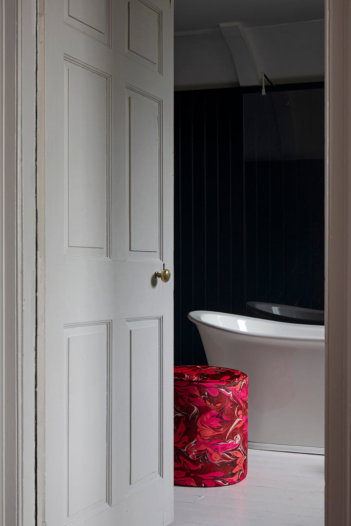A white panelled interior door is open, revealing a modern bathroom with a white freestanding bathtub and a red velvet stool covered in a vivid pink, red, and orange abstract floral pattern against a dark navy wall.