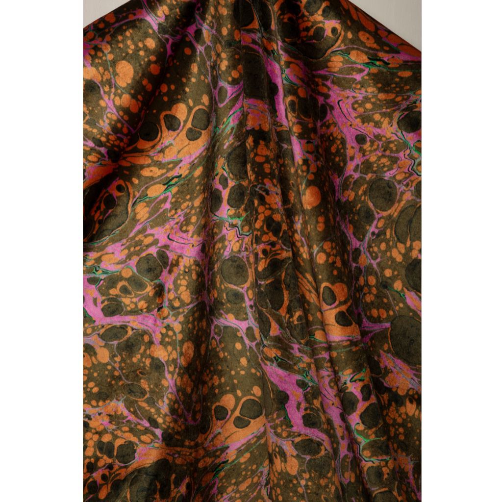 A close-up of draped fabric with an abstract marbled pattern in dark olive green, burnt orange, and bright pink, with scattered black spots.